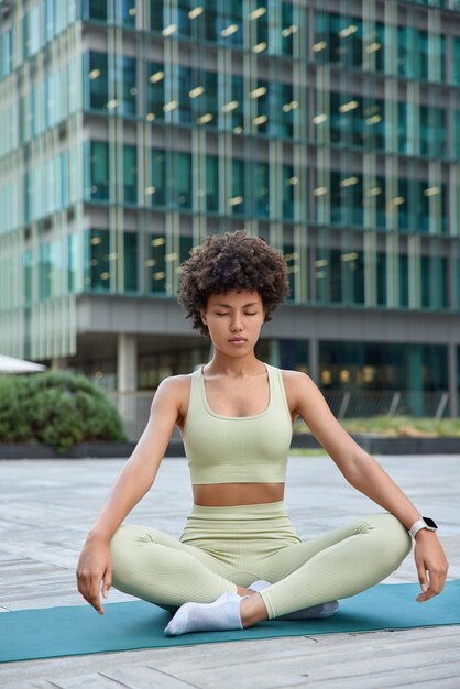 Une femme en bonne santé est assise dans une pose de lotus pratique le yoga en milieu urbain porte des vêtements de sport trouve l'équilibre respire profondément garde les yeux fermés. Un instructeur de fitness motivé s'étire avant le Pilates pendant la journée