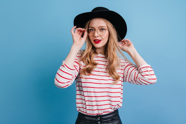 Femme bouclée au chapeau élégant regardant la caméra. Photo de Studio d'une fille blonde fascinante en chemise rayée.