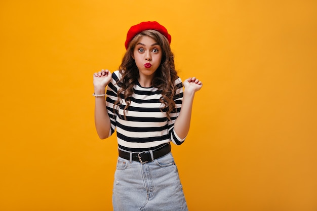 Femme bouclée de bonne humeur fait la grimace. Fille élégante en béret rouge et vêtements à la mode s'amuse sur fond isolé.