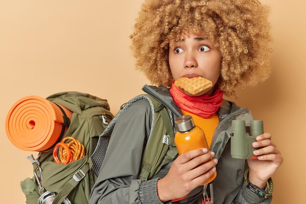 Une femme bouclée effrayée inquiète pose avec une délicieuse gaufre maison dans la bouche tient des jumelles et une bouteille avec de l'eau porte un sac à dos a une randonnée isolée sur fond marron Concept de loisirs