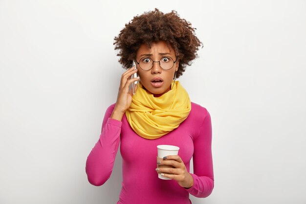 Femme bouclée embarrassée parle via smartphone, entend de mauvaises informations, fait un appel téléphonique, tient du café à emporter, regarde à travers des lunettes optiques, col polo et écharpe, isolé sur fond blanc