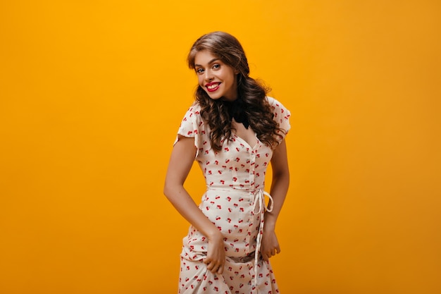 Femme bouclée en jolie robe souriant sur fond orange. Jeune fille aux cheveux ondulés et aux lèvres brillantes dans des vêtements modernes souriant.