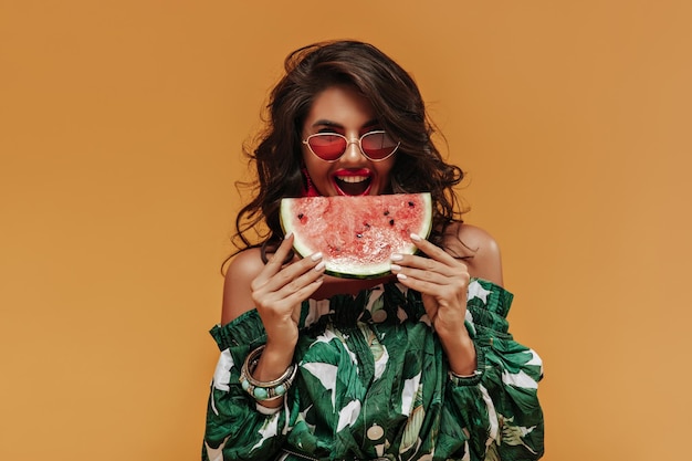 Femme bouclée joyeuse de bonne humeur dans des vêtements verts élégants et des lunettes de soleil rouges regardant dans la caméra et mangeant de la pastèque