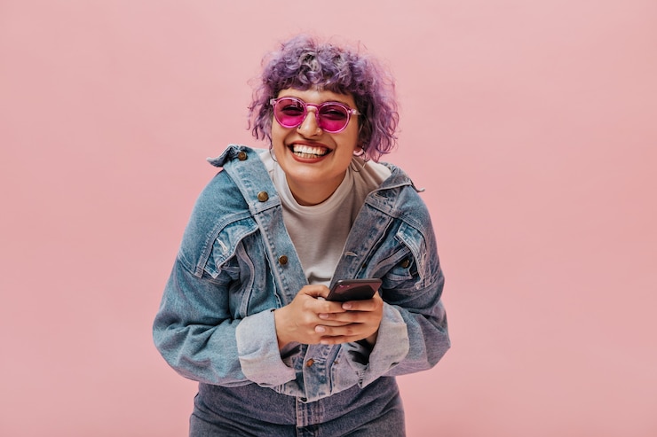 Photo gratuit femme bouclée lumineuse aux cheveux lilas dans des lunettes roses élégantes, une veste en jean et un jean tient son smartphone