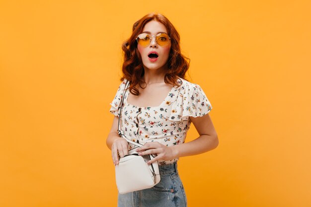 Femme bouclée à lunettes de soleil regarde confusément la caméra et tient un petit sac blanc.