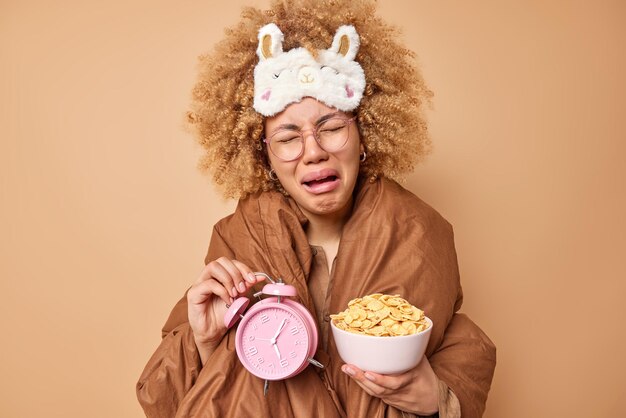 Une femme bouclée qui pleure mécontente a une expression boudeuse tient un bol de réveil de cornflakes se réveille tôt le matin porte un masque de sommeil sur le front enveloppé dans une couette isolée sur fond marron