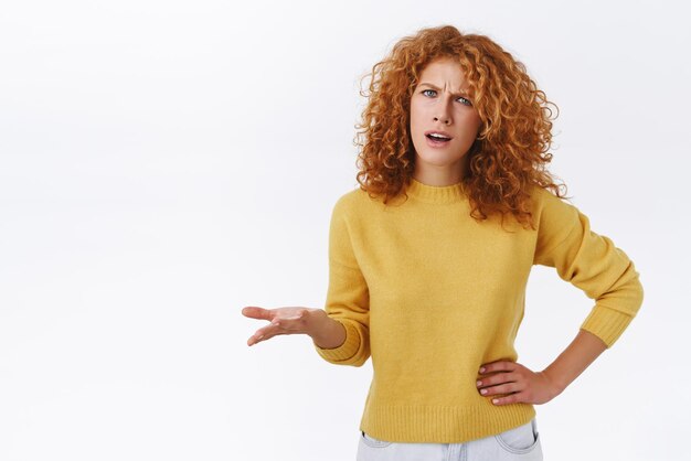 Femme bouclée rousse interrogée frustrée en pull jaune ayant un argument incompréhension fronçant les sourcils lever une main dans la consternation regardant dérangé et perplexe se plaindre fond blanc