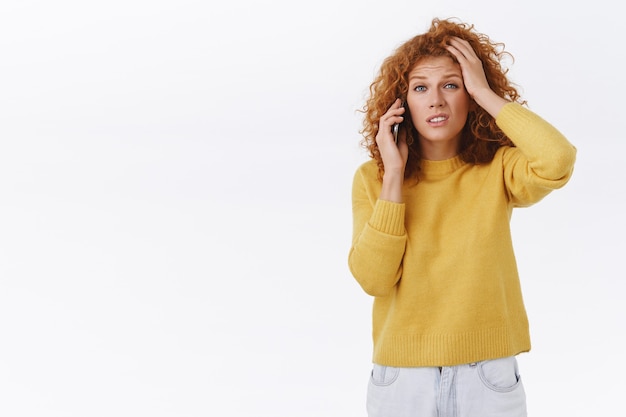 Femme bouclée rousse perplexe et indécise en pull jaune, tête tactile, fronçant les sourcils, grimaçant perplexe, parlant au téléphone mobile