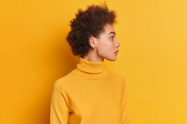 femme bouclée se tient sur le côté, a une expression sérieuse cheveux bouclés foncés vêtus de col roulé porte des boucles d'oreilles rondes