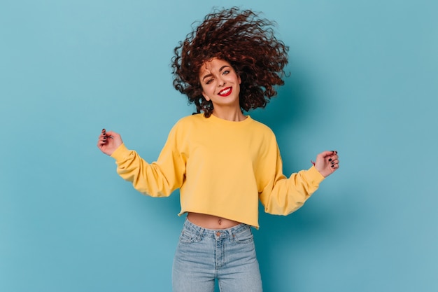 Femme bouclée souriante aux lèvres rouges vêtue d'un pull jaune et d'un jean skinny jouant les cheveux sur un espace isolé.