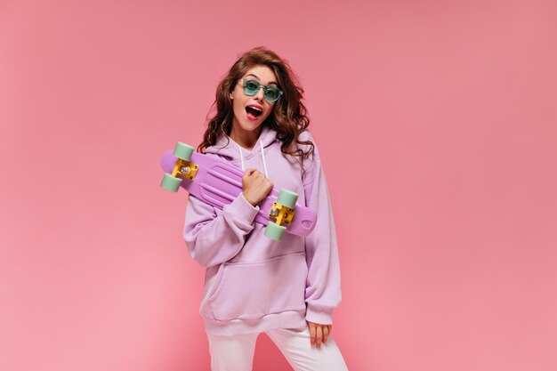 Femme bouclée surprise en sweat à capuche surdimensionné violet tient un longboard coloré Charmante dame en tenue sportive et lunettes de soleil vertes pose sur fond rose isolé