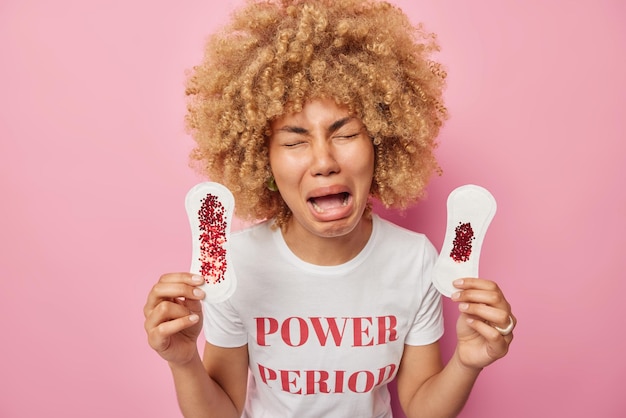 Une femme bouleversée a de la mauvaise humeur pendant la menstruation tient deux serviettes hygiéniques sales a une expression lugubre pleure de désespoir vêtue d'un t-shirt décontracté isolé sur fond rose Hygiène intime