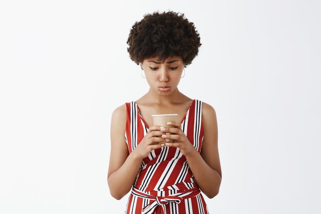 Femme bouleversée et sombre avec une coiffure afro et une peau foncée se pincant les lèvres et regardant avec tristesse une tasse de papier vide de café voulant une recharge, posant