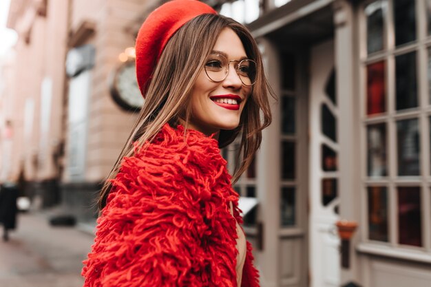 Femme bronzée aux cheveux noirs avec rouge à lèvres posant sur la rue. Portrait de jeune femme élégante en manteau tricoté rouge et bonnet.