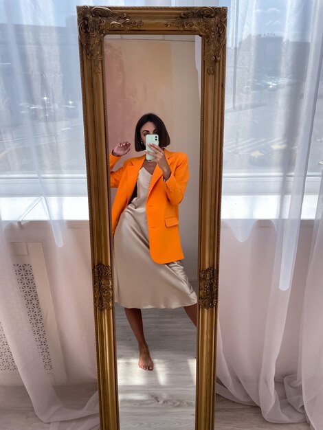 Une femme bronzée en forme dans une robe en soie beige romantique et un blazer orange à la maison prend un selfie photo au téléphone dans un miroir