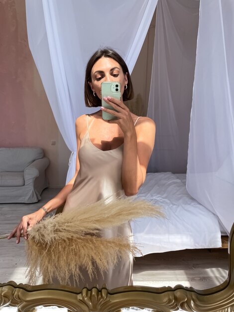 Une femme bronzée en forme en robe de soie beige romantique à la maison prend un selfie photo au téléphone dans un miroir