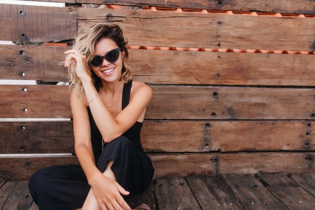 Femme bronzée à lunettes de soleil assis les jambes croisées. magnifique jeune femme en tenue noire posant sur un mur en bois.