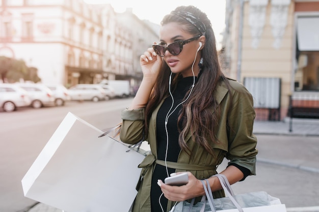 Femme bronzée sérieuse dans des lunettes noires écoutant de la musique dans des écouteurs blancs. Charmante dame hispanique en veste tendance portant des achats, marchant dans la rue.