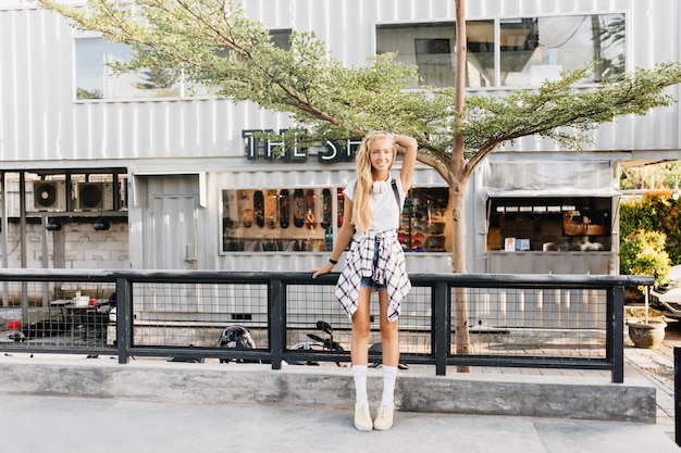 femme bronzée souriante aux cheveux blonds posant sur fond de rue.