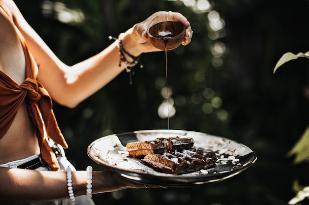 Femme bronzée en soutien-gorge brun détient un bol avec du sirop d'érable et pose dans le jardin