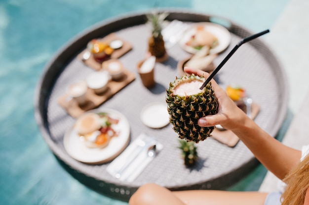 Photo gratuite femme bronzée tenant un cocktail d'ananas sucré. modèle féminin posant pendant le déjeuner dans la piscine.