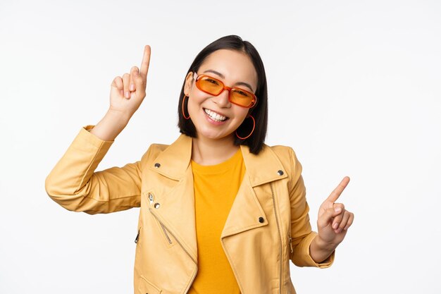 Femme brune asiatique souriante à lunettes de soleil pointant les doigts sur le côté gauche et à droite montrant des variantes riant et dansant portant des lunettes de soleil fond blanc