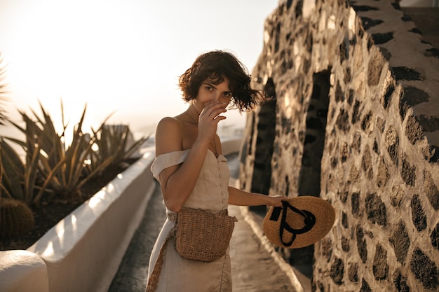 Photo gratuite femme brune aux yeux bruns en robe beige avec sac de paille tient un chapeau à larges bords et se déplace le long du chemin dans la vieille ville