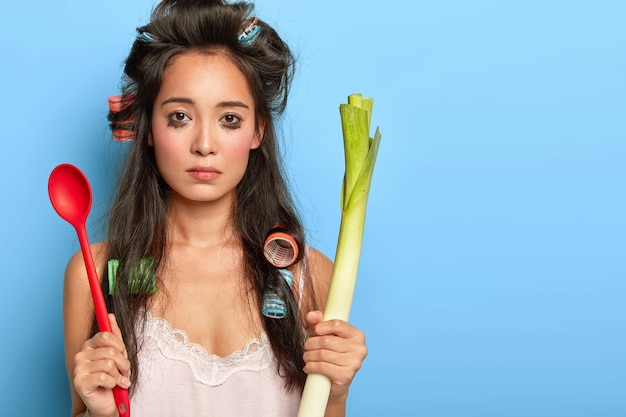 Une femme brune bouleversée pleure car elle a beaucoup de travail à la maison, ne peut pas préparer quelque chose de délicieux, a gâté le maquillage, tient le poireau vert et la cuillère n'a pas d'humeur pour la cuisine