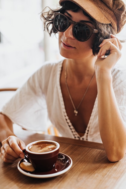 Femme brune bronzée en casquette marron et lunettes de soleil détient une tasse en porcelaine brune avec café et repos au café