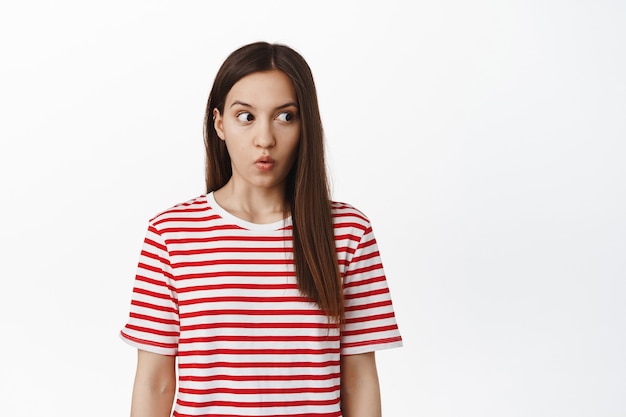 Une femme brune choquée haletant, regarde droit dans l'espace vide avec une expression surprise sans voix, vérifiant quelque chose d'impressionnant, debout contre un mur blanc.