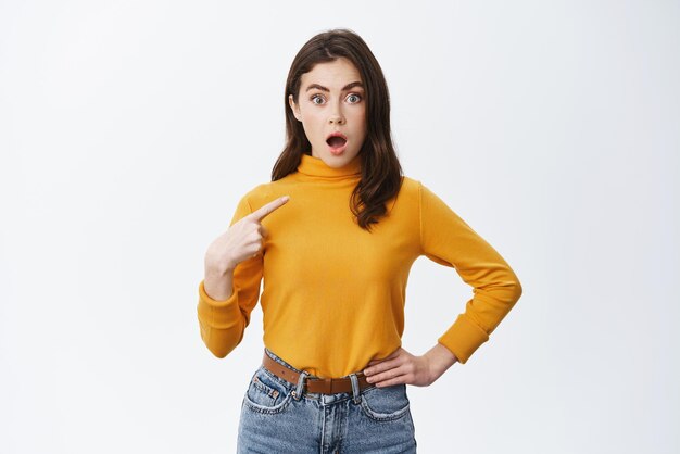 Femme brune choquée pointant vers elle-même avec la bouche ouverte regardant avec incrédulité debout sur fond blanc