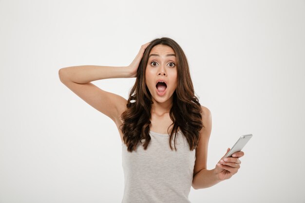 Femme brune choquée avec smartphone tenant la tête et regardant la caméra sur gris