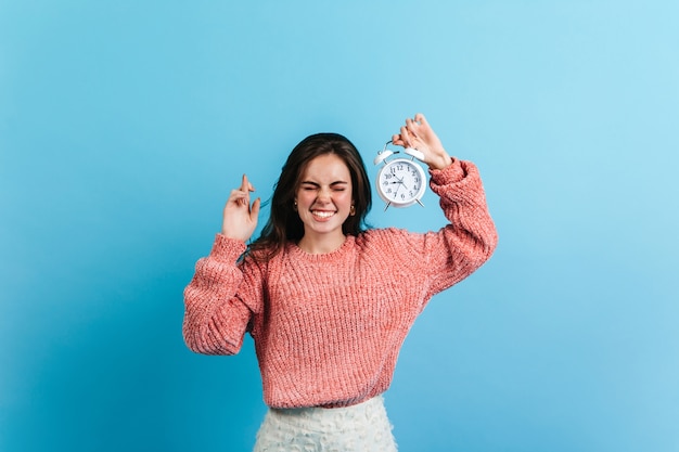 Femme brune a croisé ses doigts et tient un réveil blanc. Modèle en pull surdimensionné posant sur un mur bleu.