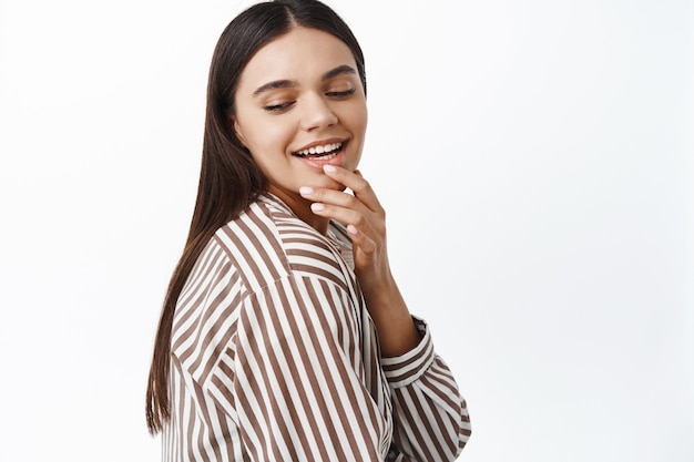 Femme brune élégante touchant ses lèvres et souriante, regardant vers le bas avec un visage coquet et séduisant, debout contre un mur blanc