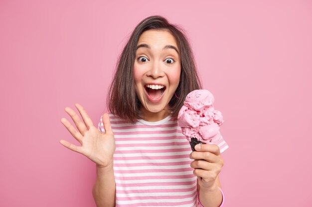 Une femme brune émotionnelle s'exclame bruyamment en gardant la paume levée mange une délicieuse crème glacée réagit à quelque chose d'inattendu a surpris un look joyeux porte un t-shirt à rayures décontracté isolé sur un mur rose