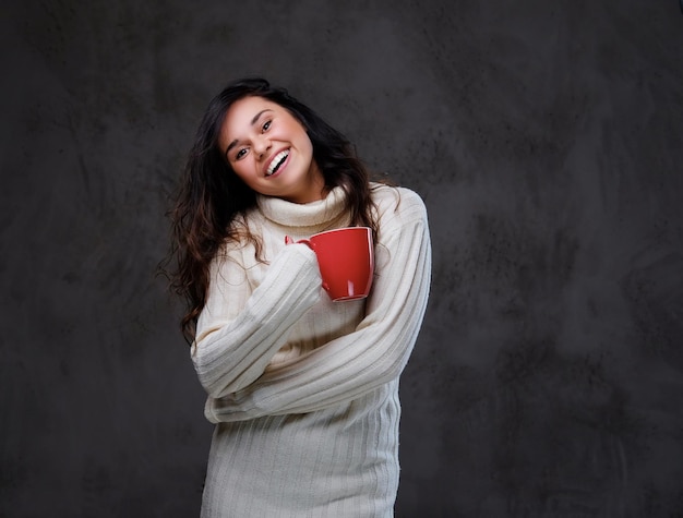 Femme brune émotionnelle et souriante vêtue d'un chandail chaud à col roulé tient une grande tasse de café rouge sur fond gris.