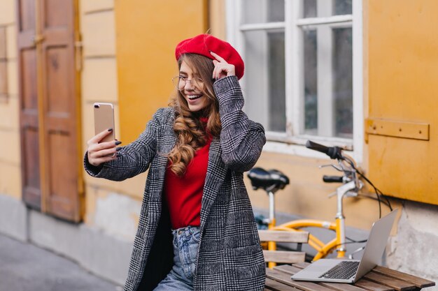 Femme brune glamour en tenue mignonne faisant un selfie dans la rue avec un vélo jaune en arrière-plan. Photo extérieure d'une fille en tenue tendance debout près d'un ordinateur portable.