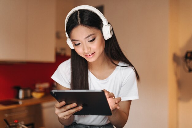 Femme brune en haut blanc et écouteurs de bonne humeur se penche sur l'écran de la tablette