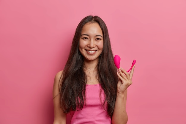 Une femme brune heureuse pose avec un vibrateur bluetooth intelligent, utilise une application spéciale sur mobile pour améliorer l'orgasme, tient un outil sexuel pour améliorer le plaisir, isolé sur un mur rose. Technologies modernes et vie sexuelle