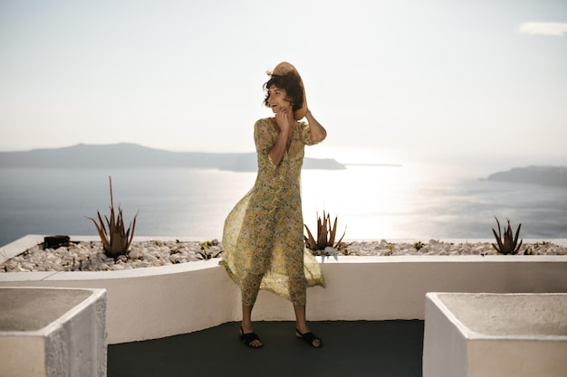 Une femme brune heureuse tient un canotier et sourit à l'extérieur Dame aux cheveux courts en chapeau et robe à fleurs pose sur une terrasse avec vue sur la mer