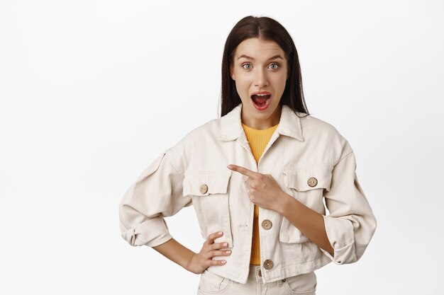 Femme brune impressionnée en veste en jean, bouche ouverte fascinée, doigt pointé à gauche de la vente, logo ou espace de copie de côté