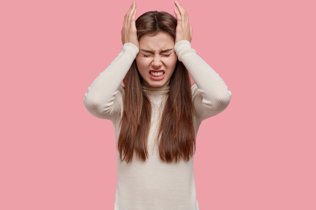 Photo gratuite femme brune inquiète épuisée garde les mains sur les tempes, serre les dents, a mal de tête ou migraine, habillée en tenue blanche, isolée sur fond rose