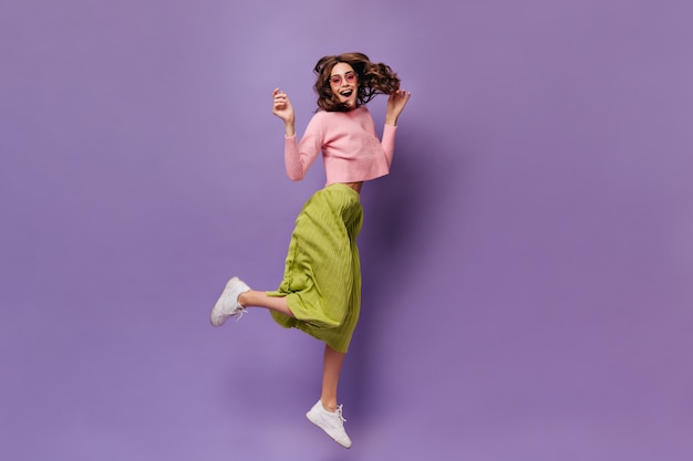 Une femme brune joyeuse en jupe verte et pull rose saute sur un mur violet
