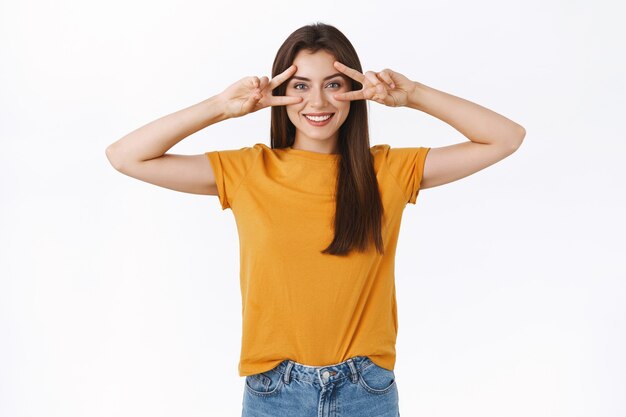 Femme brune joyeuse et optimiste en t-shirt jaune, montrant un signe de paix ou de victoire sur les yeux, souriante, exprimant la positivité et la joie, profitant de la fête, comme assister à un événement incroyable, fond blanc