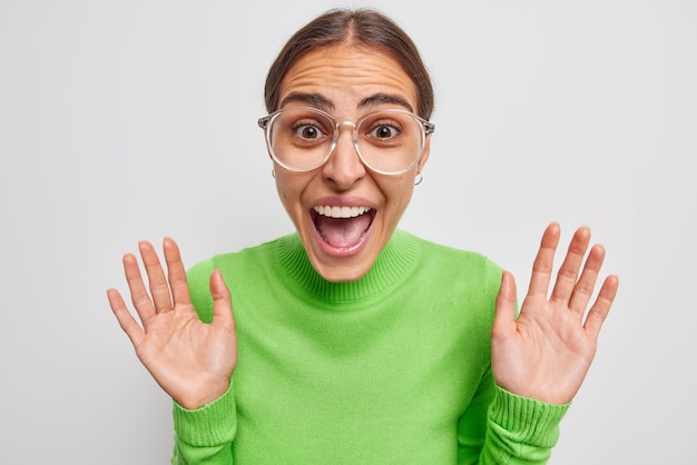 Photo gratuite une femme brune joyeuse s'exclame bruyamment se sent heureuse de réagir sur de bonnes ews garde les paumes levées porte des lunettes transparentes et un pull vert décontracté isolé sur fond blanc wow c'est incroyable