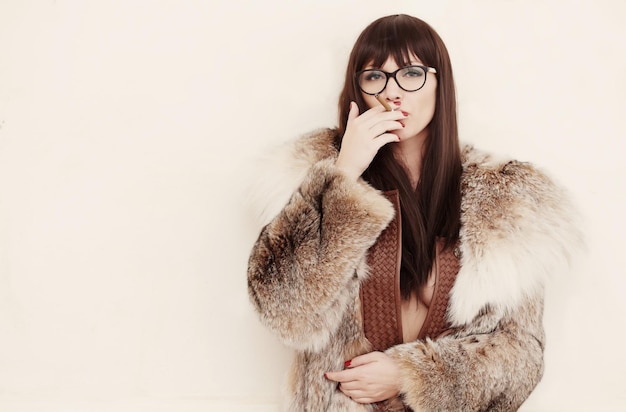 Femme brune à lunettes et dans un manteau de fourrure fumant une cigarette.