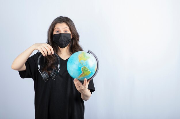 Femme brune en masque médical tenant le globe terrestre avec des écouteurs.