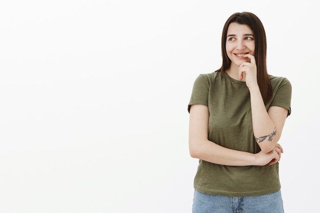 Femme brune ordinaire mignonne rêveuse et sensuelle avec tatouage et yeux bruns souriant tendrement et regardant le coin supérieur gauche avec un sourire sincère et romantique ravi, touchant la lèvre timide sur un mur gris