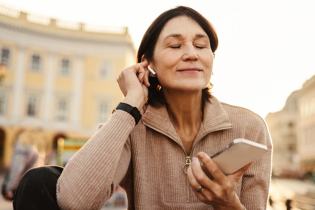 Une femme brune à la peau claire adulte pacifiée, les yeux fermés, écoute de la musique via des écouteurs dans la rue Mode technologie et concept de personnes