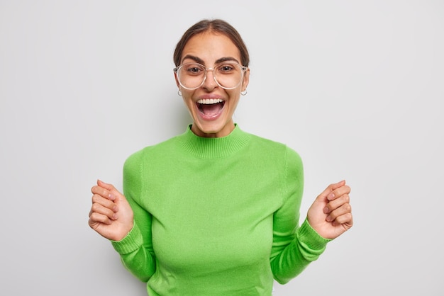 Une femme brune positive lève les mains s'exclame bruyamment porte des lunettes transparentes et un cavalier vert décontracté se sent très heureux entend des nouvelles positives isolées sur fond blanc Concept d'émotions